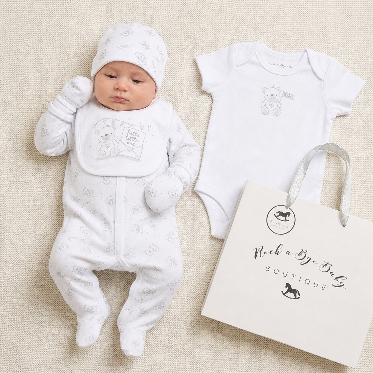 Newborn baby in white outfit with matching hat and body suit, next to a 'Rock a Bye Baby Boutique' bag on a beige background.