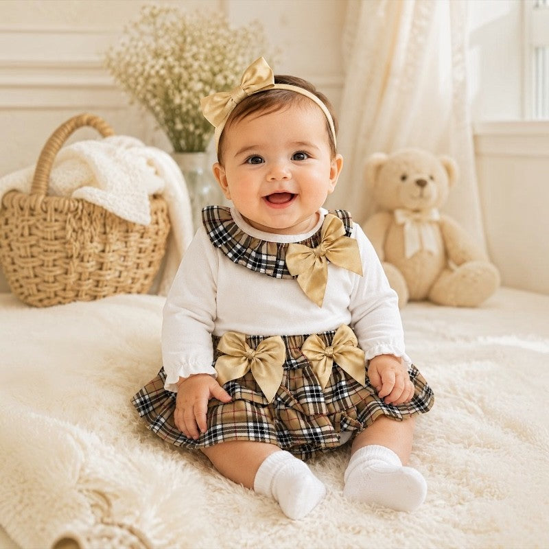 A baby girl sitting on a white fluffy blanket wearing a white long-sleeve top, a brown and beige plaid skirt with a large beige bow at the waist, and a matching beige bow headband. A woven basket, white flowers, and a teddy bear are in the background.