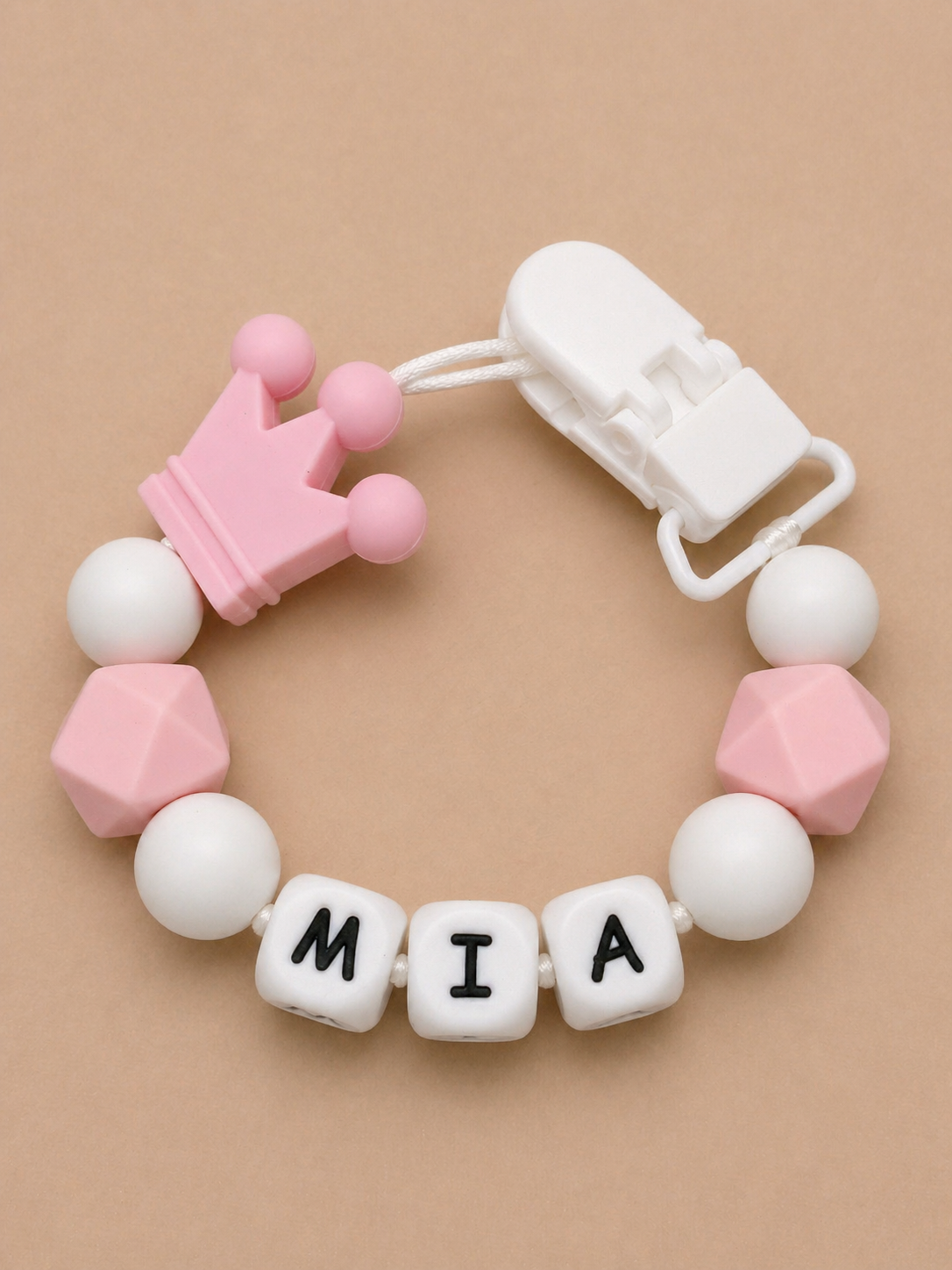 Baby teether with pink crown and letter blocks spelling 'MIA' on a beige background