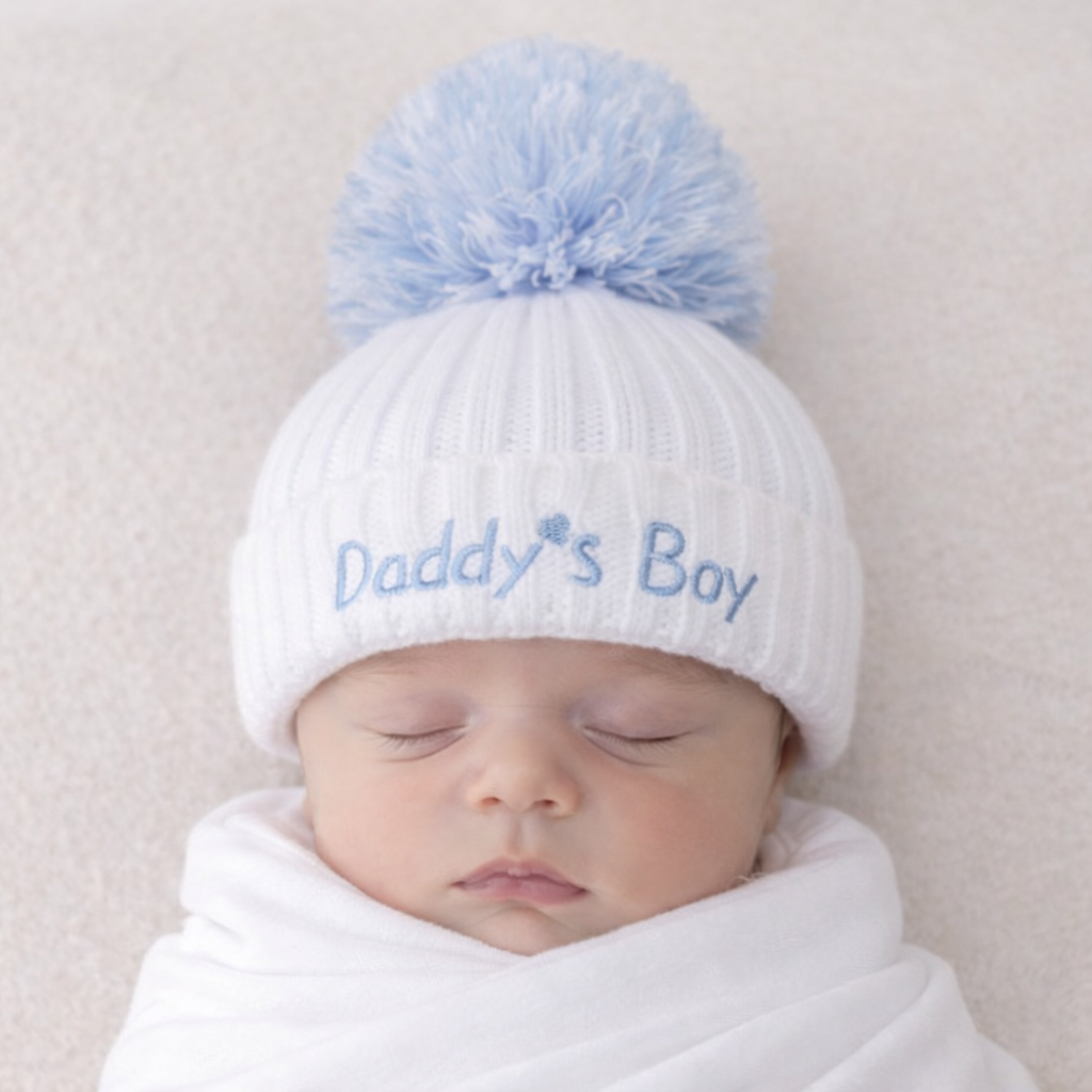 Newborn baby wearing a white knit hat with a blue pom-pom and 'Daddy's Boy' text on a light background.