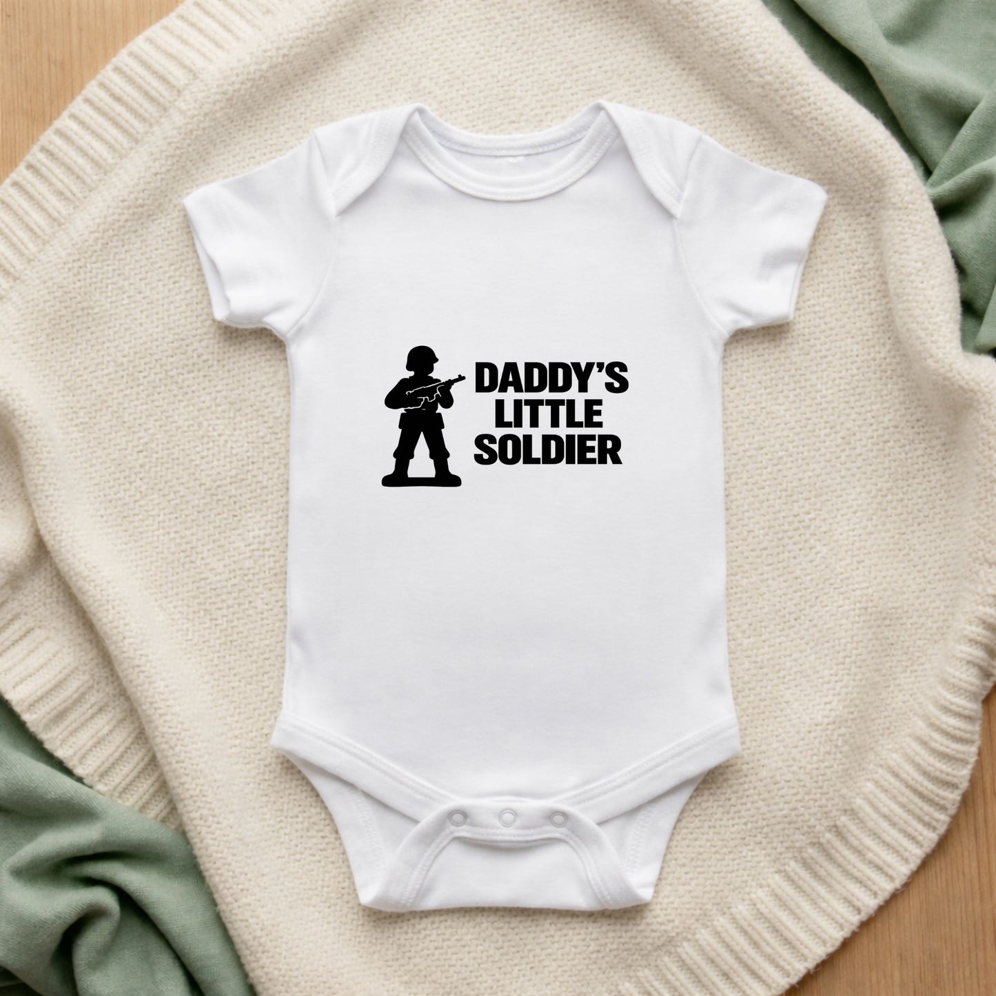 White baby onesie with 'Daddy's Little Soldier' text and graphic on a textured surface.
