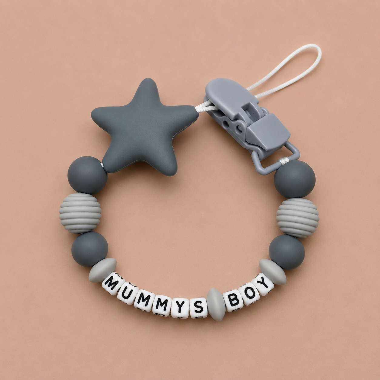 Silicone dummy clip with star-shaped clip and 'MUMMYS BOY' beads on a beige background