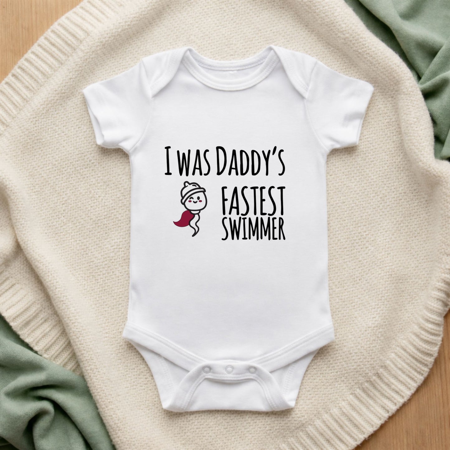 White baby onesie with text 'I was daddy's fastest swimmer' on a textured surface.