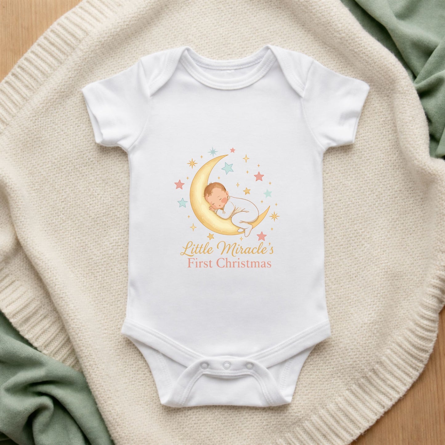 White baby onesie with 'Little Miracle's First Christmas' design on a textured surface.