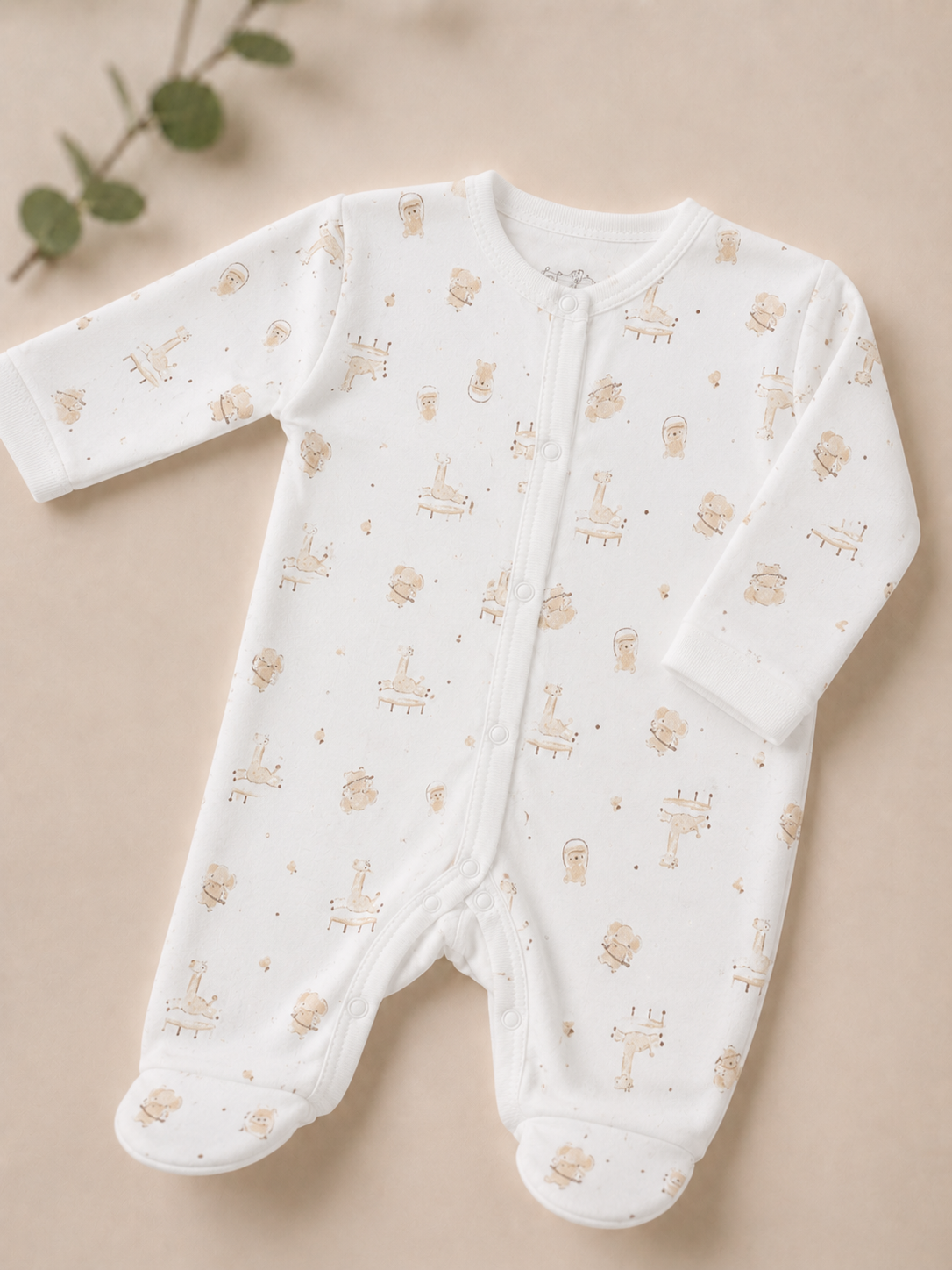 A white long-sleeve baby footed sleeper with a delicate beige and gold animal print, laid flat on a neutral beige background with a small green leaf sprig in the corner.