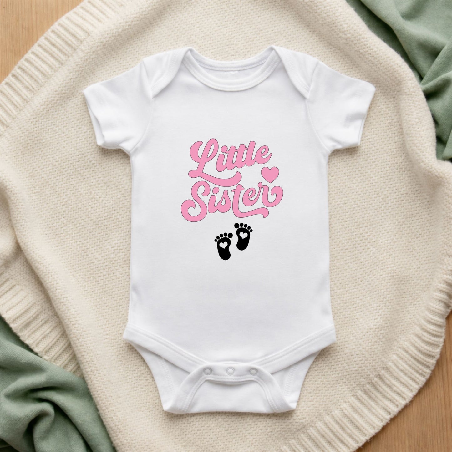 White baby onesie with 'Little Sister' in pink text and footprints on a beige surface.