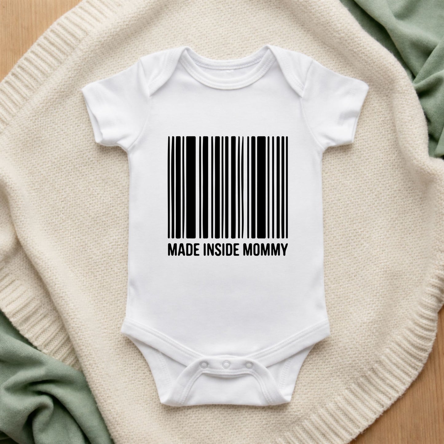 White baby onesie with a barcode and 'MADE INSIDE MOMMY' text on a textured surface.