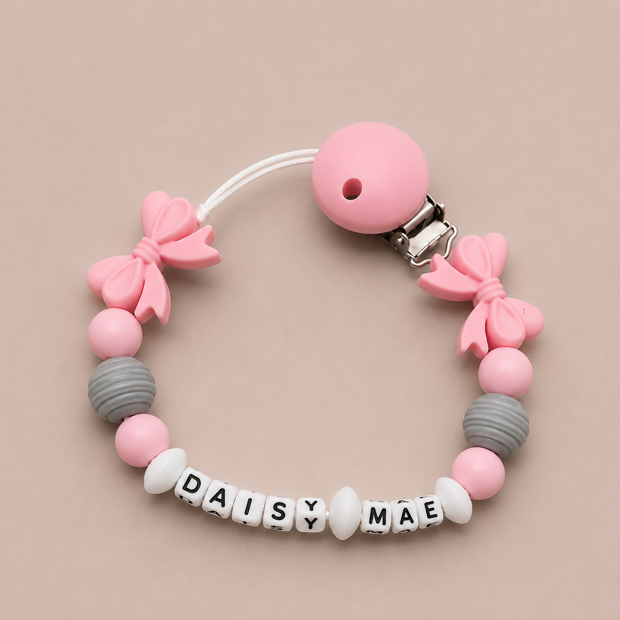 Pink and gray dummy clip with 'DAISY MAE' beads on a beige background