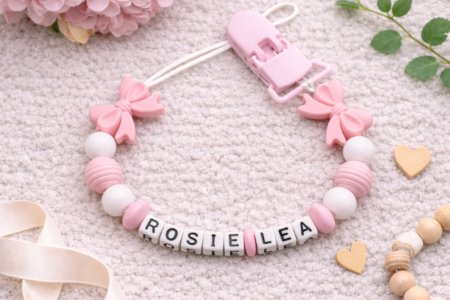 Personalised baby dummy clip with pink bows and white beads spelling “Princess” displayed on a wooden stand against a soft grey velvet background.