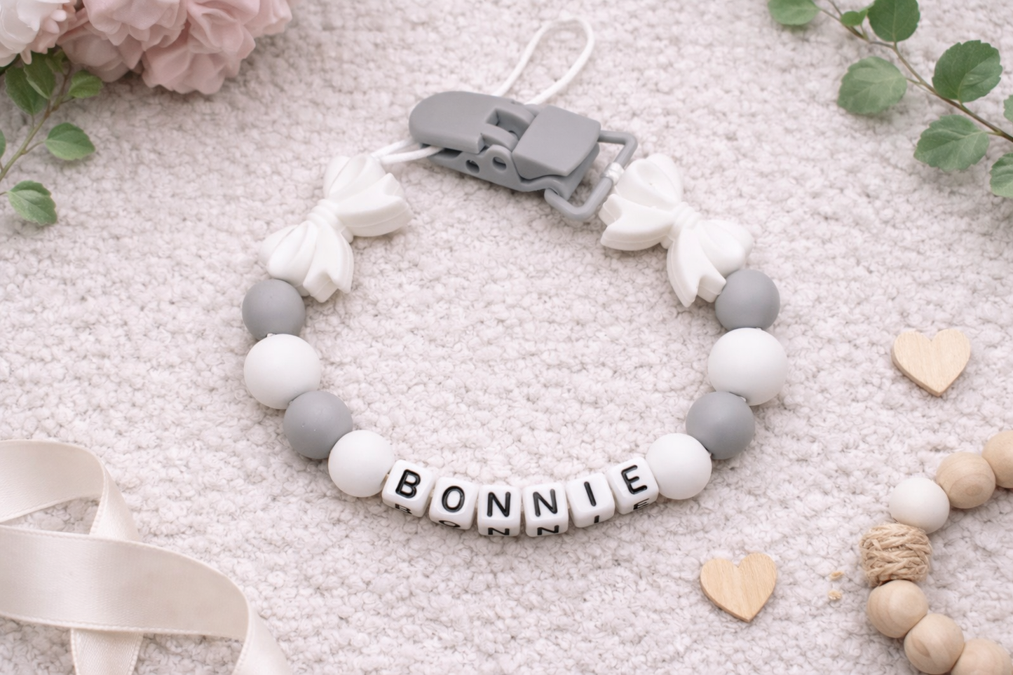 Personalized baby pacifier clip with white bow accents and grey silicone beads spelling the name Bonnie on a soft cream textured background with floral details.