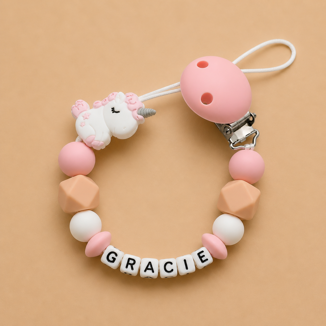 Pink and beige dummy clip with a unicorn charm and 'GRACIE' beads on a beige background