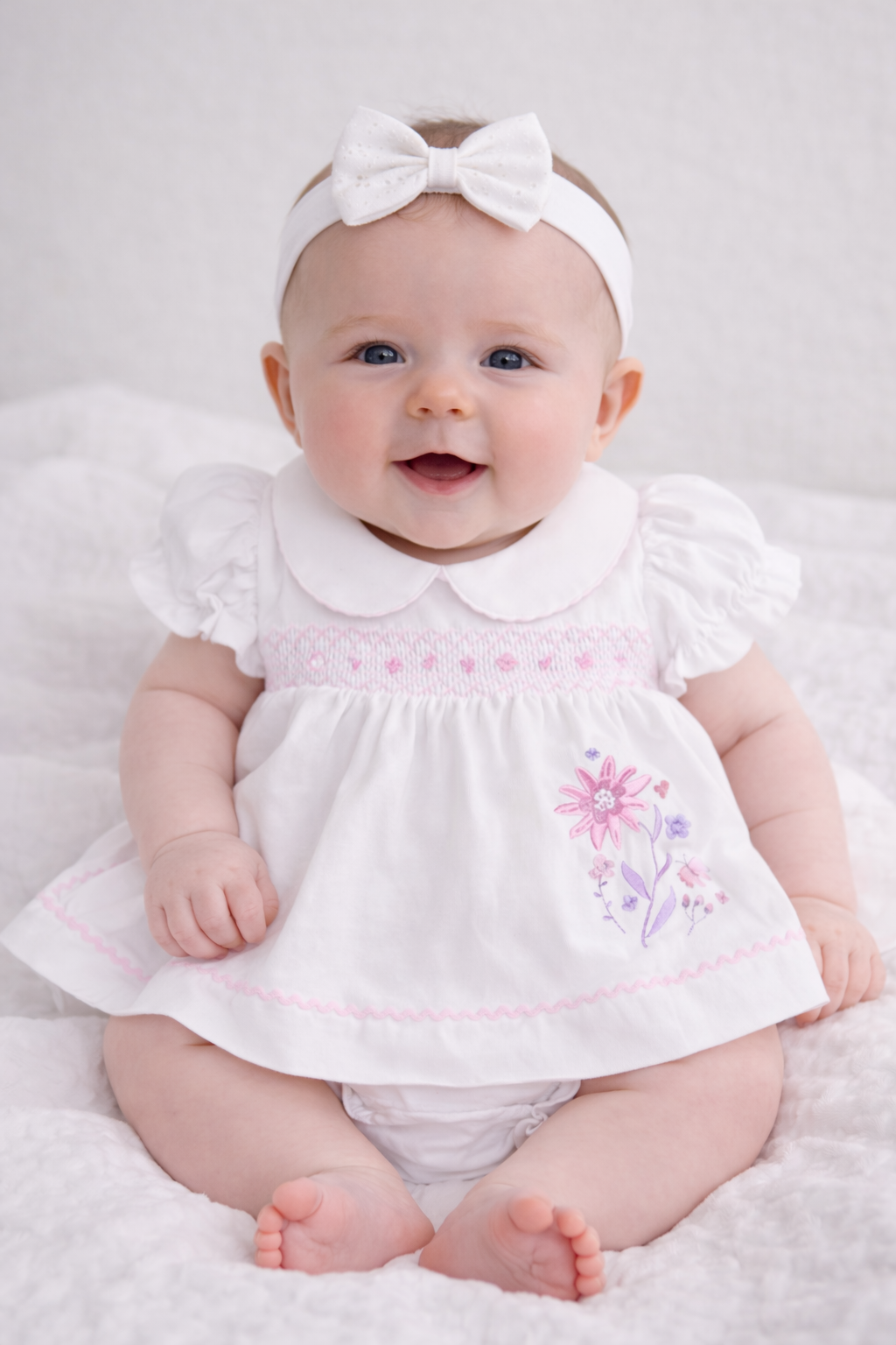 Baby wearing a white dress with floral embroidery and a matching headband on a white background