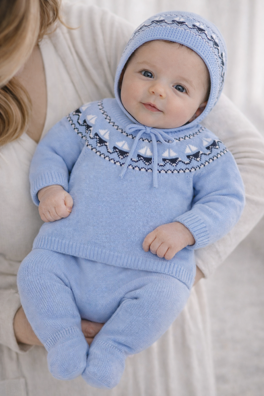 Baby wearing a blue knitted outfit with a hood, sitting on a white surface.