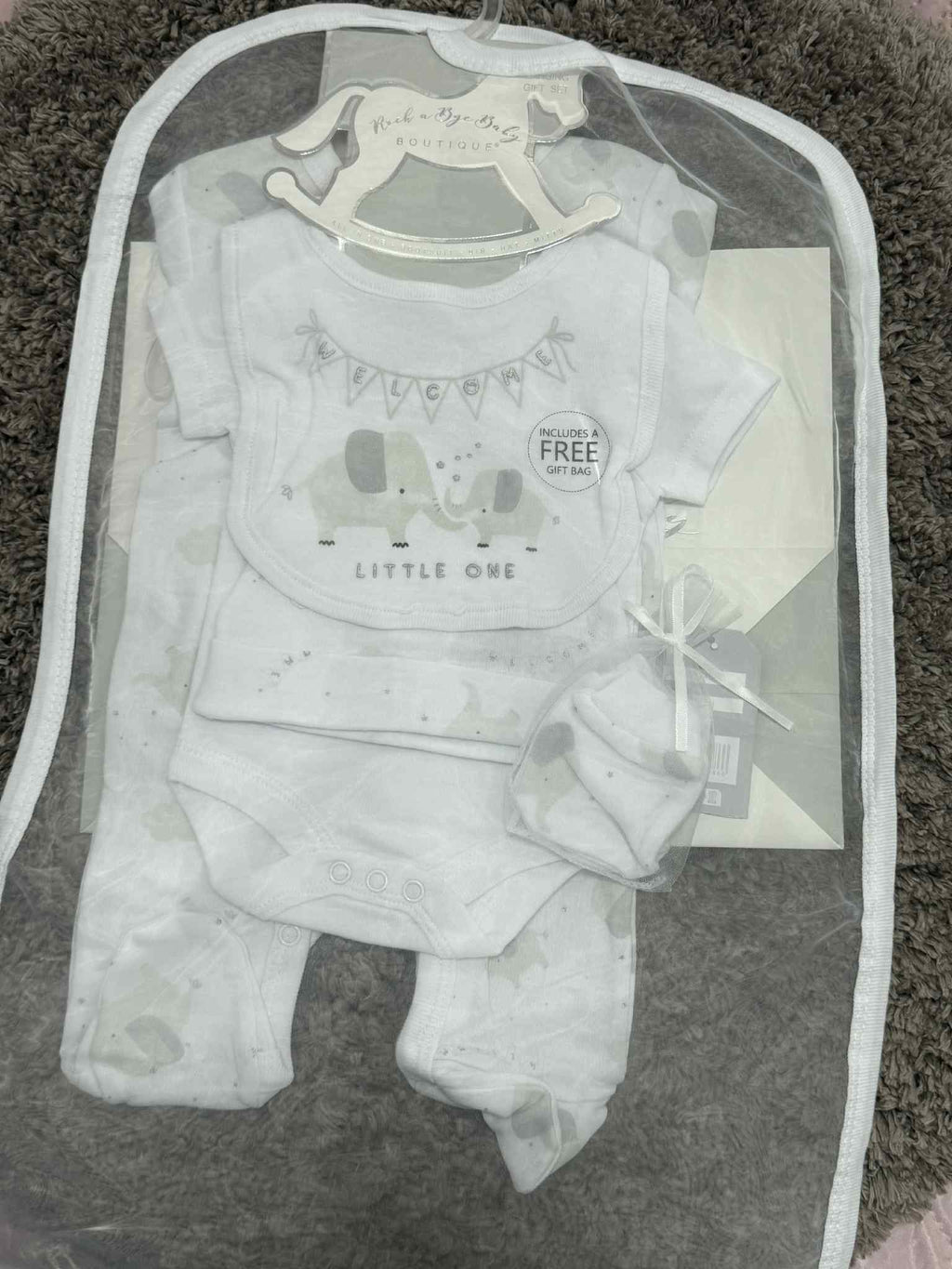 Baby outfit set with onesie, pants, and bib in a clear bag on a carpeted floor.