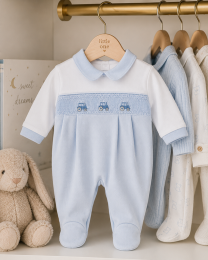 Baby blue onesie with embroidered details on a wooden hanger, surrounded by other baby clothes and a plush toy.