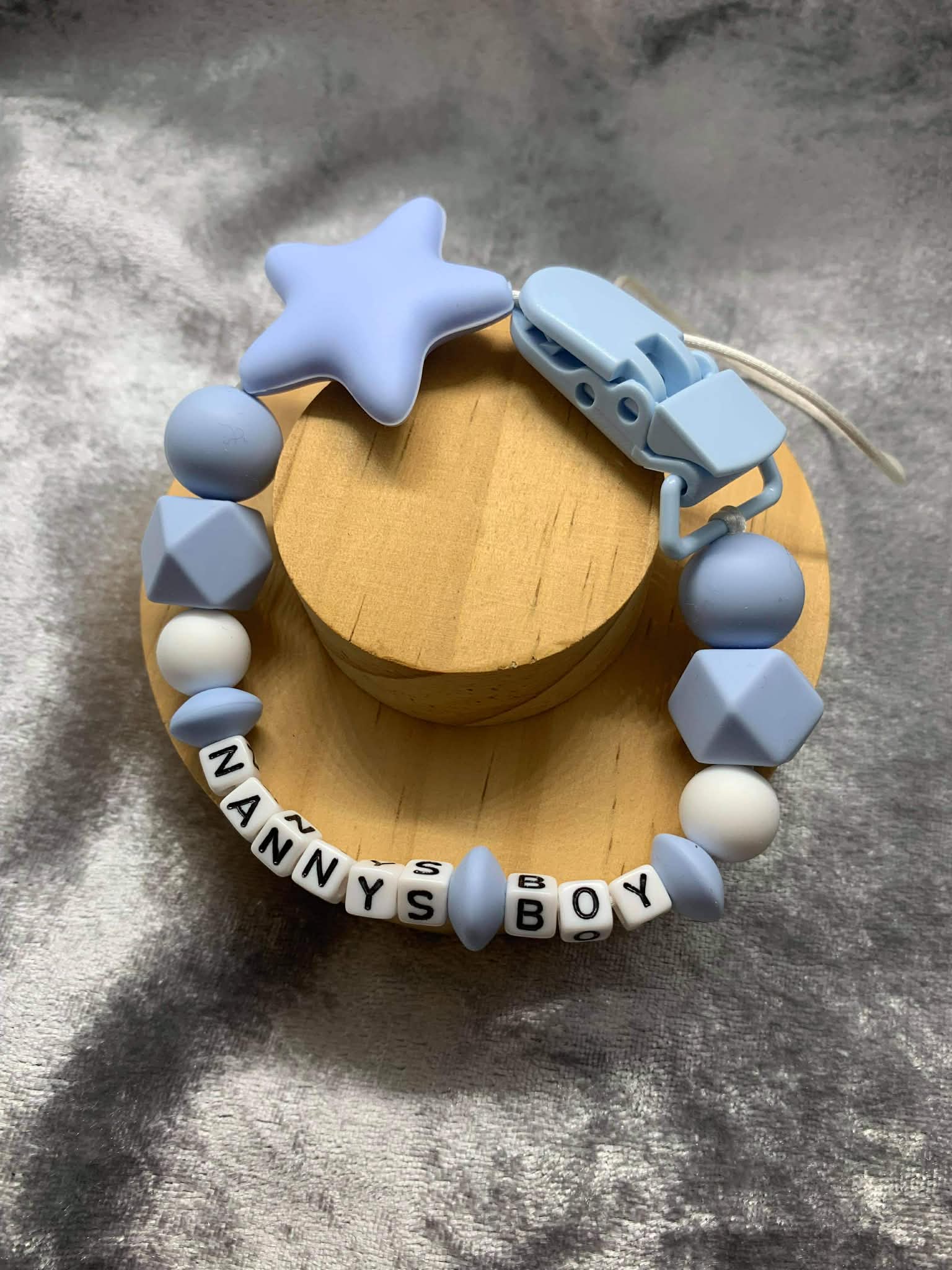 baby blue and white Silicone Dummy Clip with blue star personalised with the name 'Nanny's-Boy' on a grey textured surface. Can be personalised