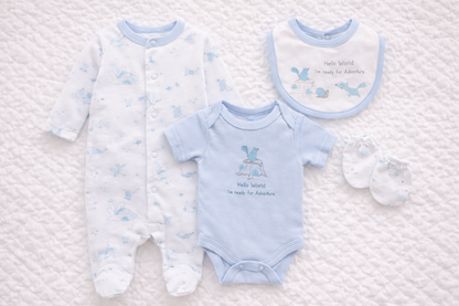 Flat lay image of a baby boys 4-piece newborn clothing set displayed on a soft white quilted background. The set includes a white footed babygrow with blue adventure animal prints, a light blue short-sleeve bodysuit with “Hello World I’m ready for Adventure” text, a matching bib with coordinating animal design, and a pair of white baby scratch mittens. Soft, coordinated newborn essentials ideal for baby showers or first outfits.