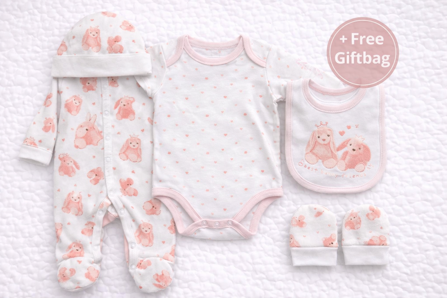 Five-piece baby girl gift set featuring a matching pink bunny print sleepsuit, short-sleeve bodysuit with heart pattern, coordinating hat, mittens, and a soft bib with bunny illustration. Beautifully styled with a + Free Giftbag, ideal for newborns and baby shower gifting.