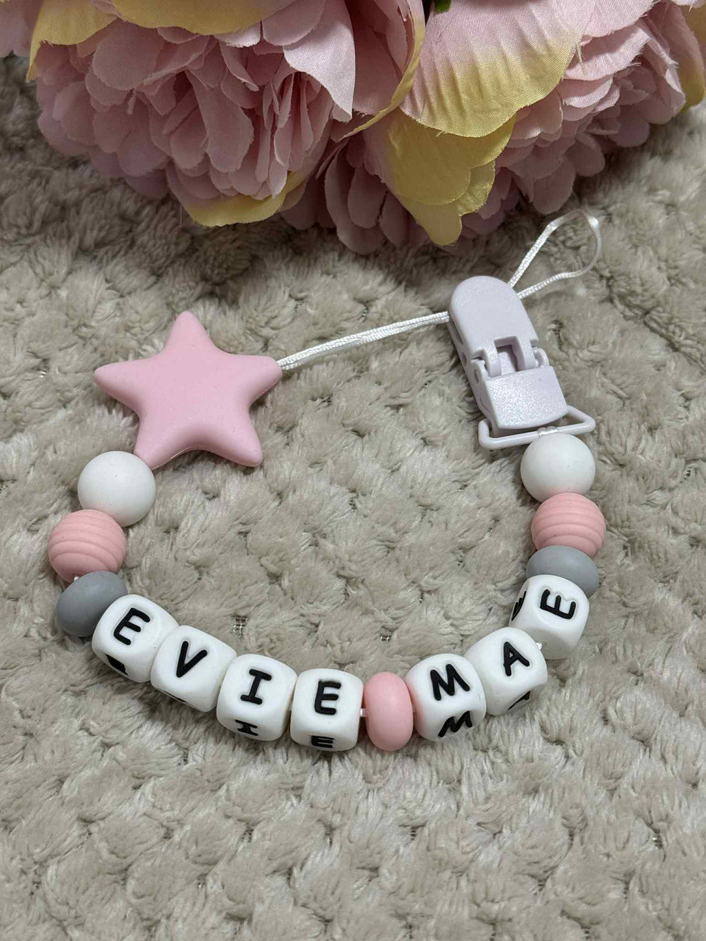 A beaded pacifier clip with white, pink, and gray beads, a pink star, and letter beads spelling "EVIE MAE," lying on a textured beige surface with pink flowers in the background.