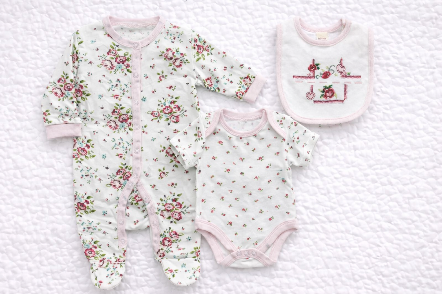 Three-piece baby girls floral outfit set featuring a long-sleeve floral babygrow, matching short-sleeve bodysuit, and coordinating embroidered bib with pink trim, displayed on a white quilted background. Soft, delicate rose prints make this set ideal for newborn wear or gifting.