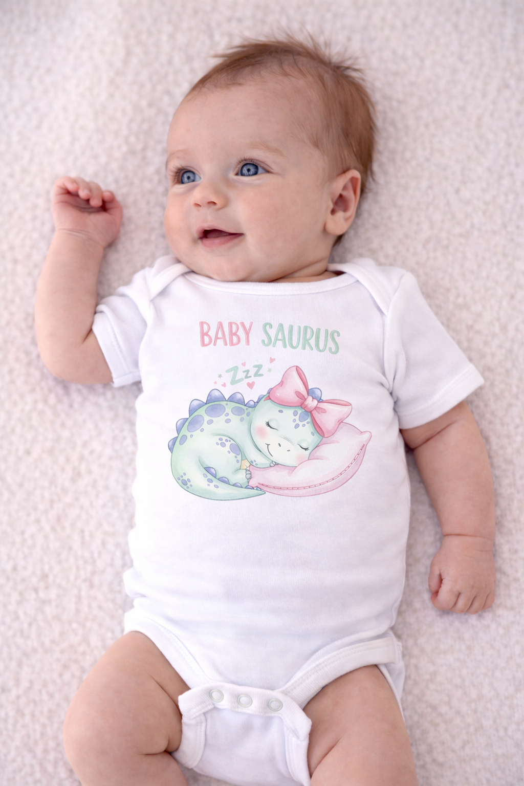 Baby wearing a white onesie with 'BABY SAURUS' print on a light pink blanket