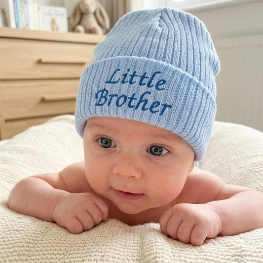 A baby lying on a cream textured blanket wearing a light blue ribbed knit beanie with 'Little Brother' embroidered in blue on the front cuff.