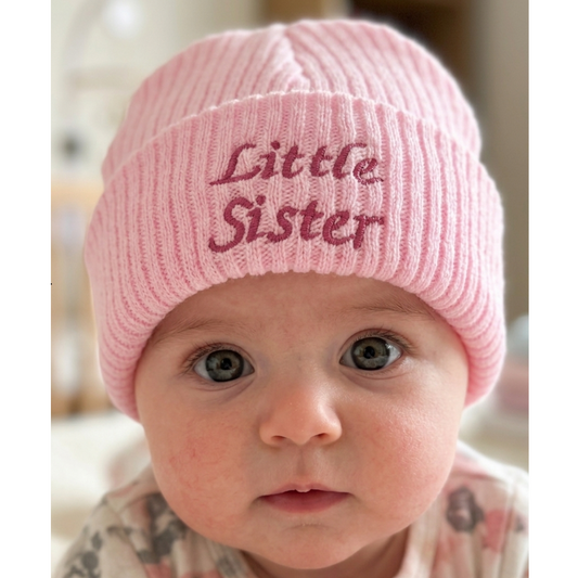 A close-up of a baby wearing a light pink ribbed knit beanie with 'Little Sister' embroidered in dark pink on the front, set against a softly blurred indoor background.