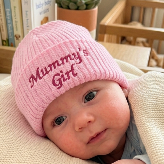A close-up of a baby wearing a pink ribbed knit beanie with 'Mummy's Girl' embroidered on the front, resting on a cream blanket in a cozy indoor setting.