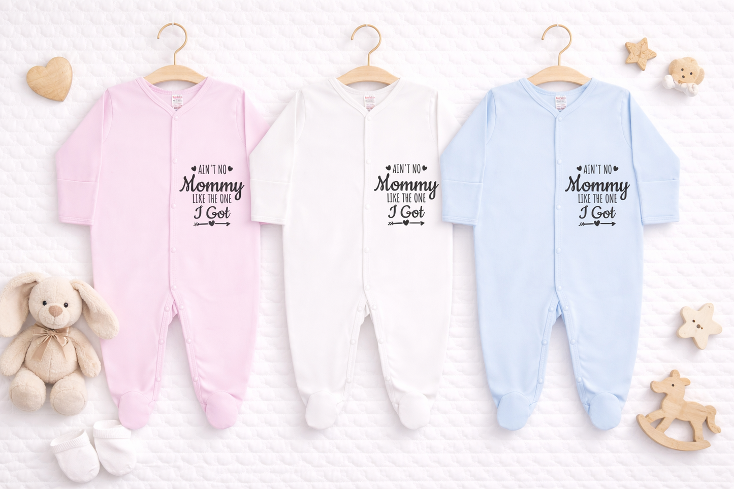 Three baby footed sleepsuits in pink, white and light blue displayed side by side on a white quilted background, each featuring the slogan “Ain’t No Mommy Like The One I Got” in black text, styled as a personalised baby gift mockup.