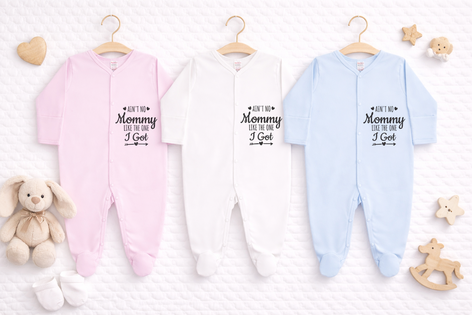 Three baby footed sleepsuits in pink, white and light blue displayed side by side on a white quilted background, each featuring the slogan “Ain’t No Mommy Like The One I Got” in black text, styled as a personalised baby gift mockup.
