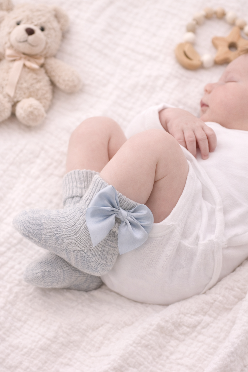 Sleeping newborn baby wearing soft grey knitted socks with satin bows, styled on a cream blanket with neutral baby toys for a cosy lifestyle mockup.