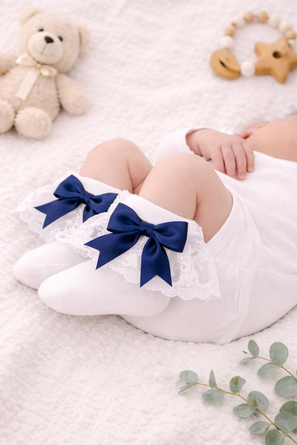 Close-up mock-up of a sleeping newborn wearing white lace baby socks finished with elegant navy blue satin bows, styled on a soft neutral blanket with classic baby accessories, ideal for product listings and gift inspiration.