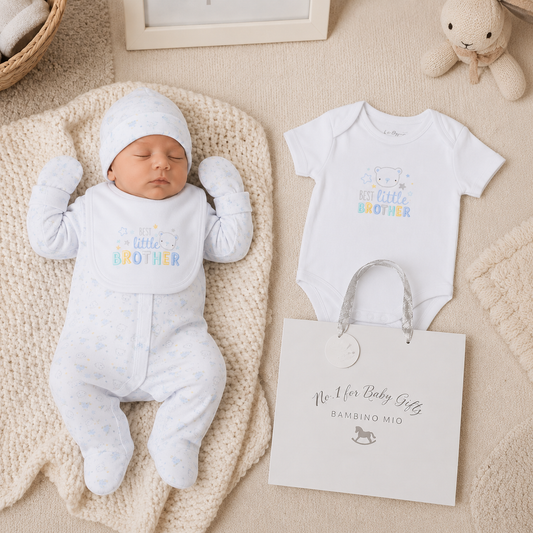 Newborn baby in a white onesie with 'little brother' text, next to a matching outfit and a gift bag on a beige blanket.