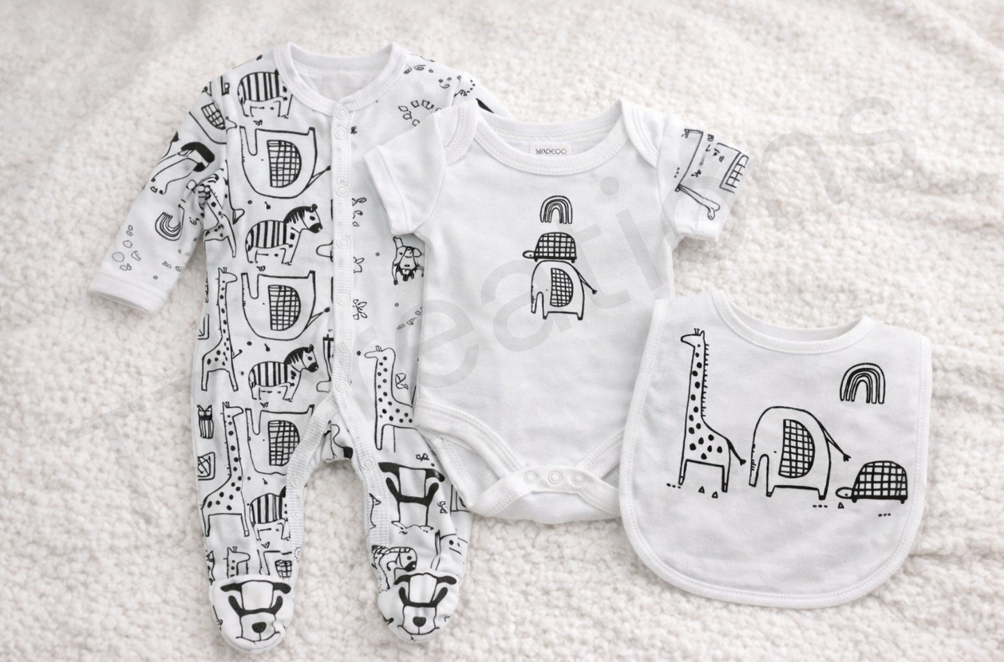 Monochrome safari-themed baby gift set featuring a patterned footed sleepsuit, short-sleeve bodysuit, and matching bib with giraffe, elephant, turtle and rainbow illustrations, laid flat on a soft cream background.