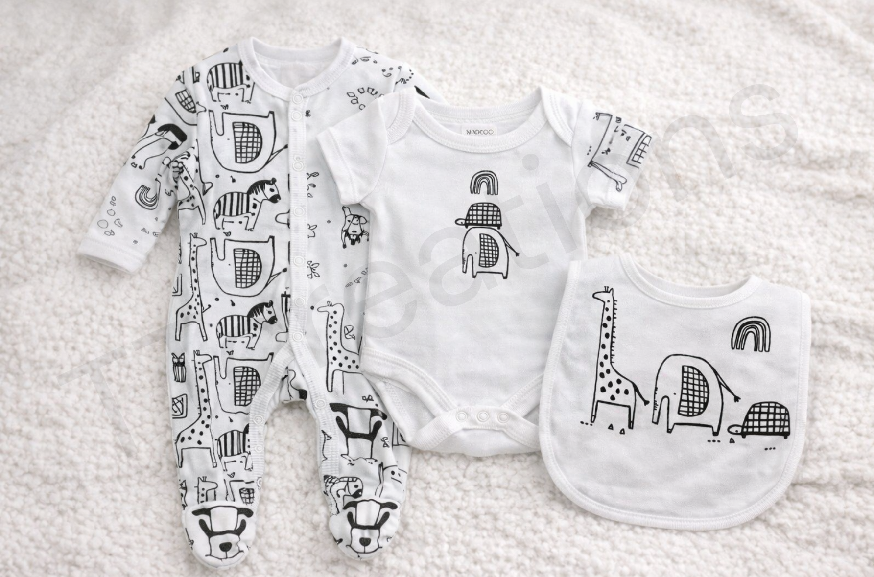 Monochrome safari-themed baby gift set featuring a patterned footed sleepsuit, short-sleeve bodysuit, and matching bib with giraffe, elephant, turtle and rainbow illustrations, laid flat on a soft cream background.