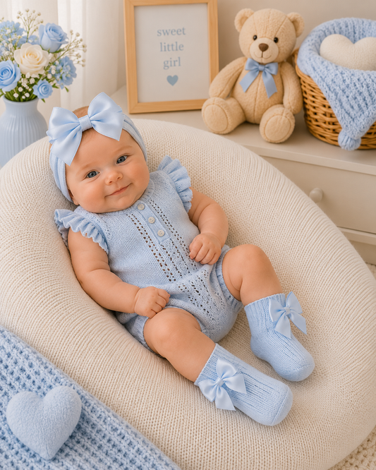Baby in blue outfit with matching headband and socks, surrounded by baby items like a teddy bear and flowers.