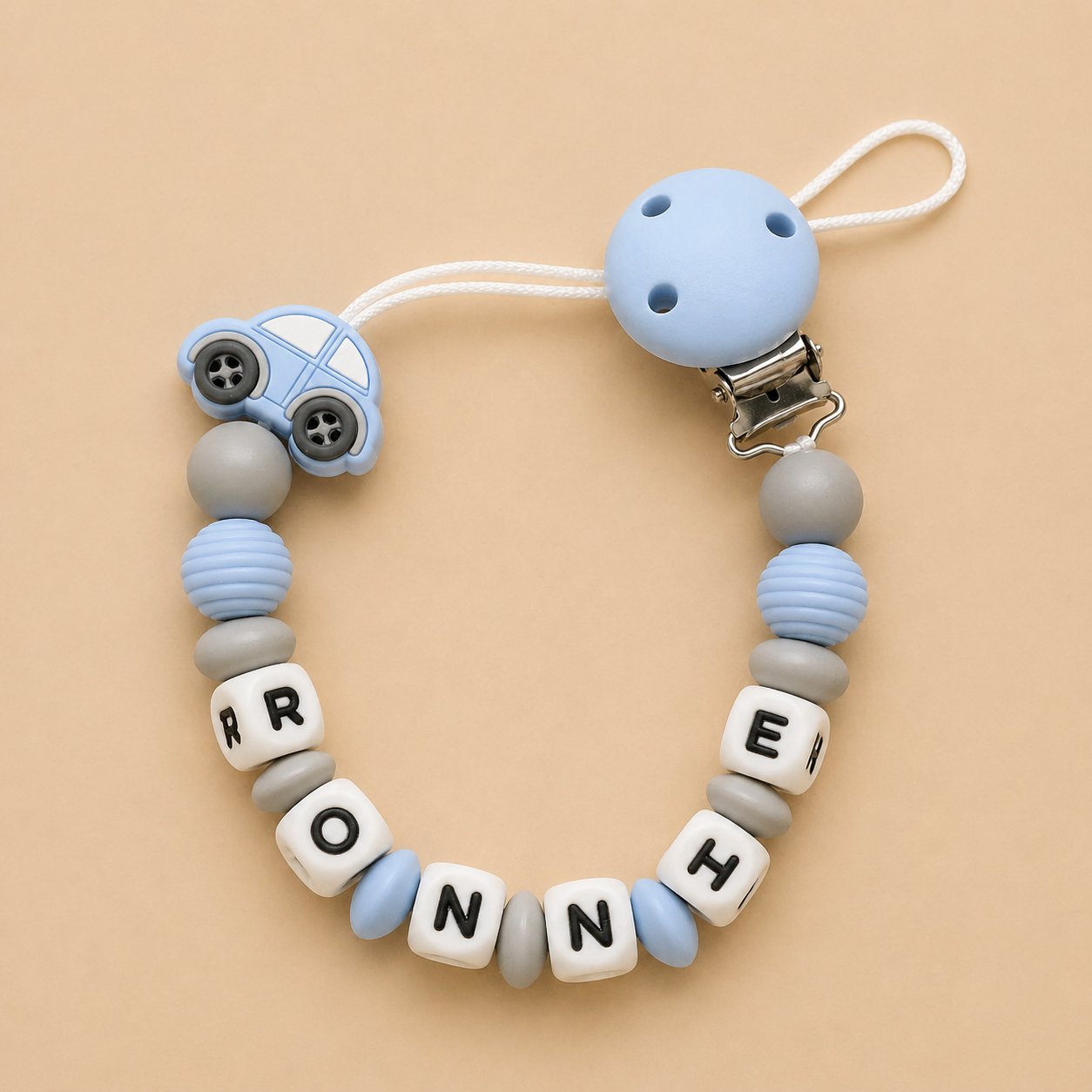 Baby dummy clip with blue car charm and letter beads spelling 'RONNIE' on a beige background