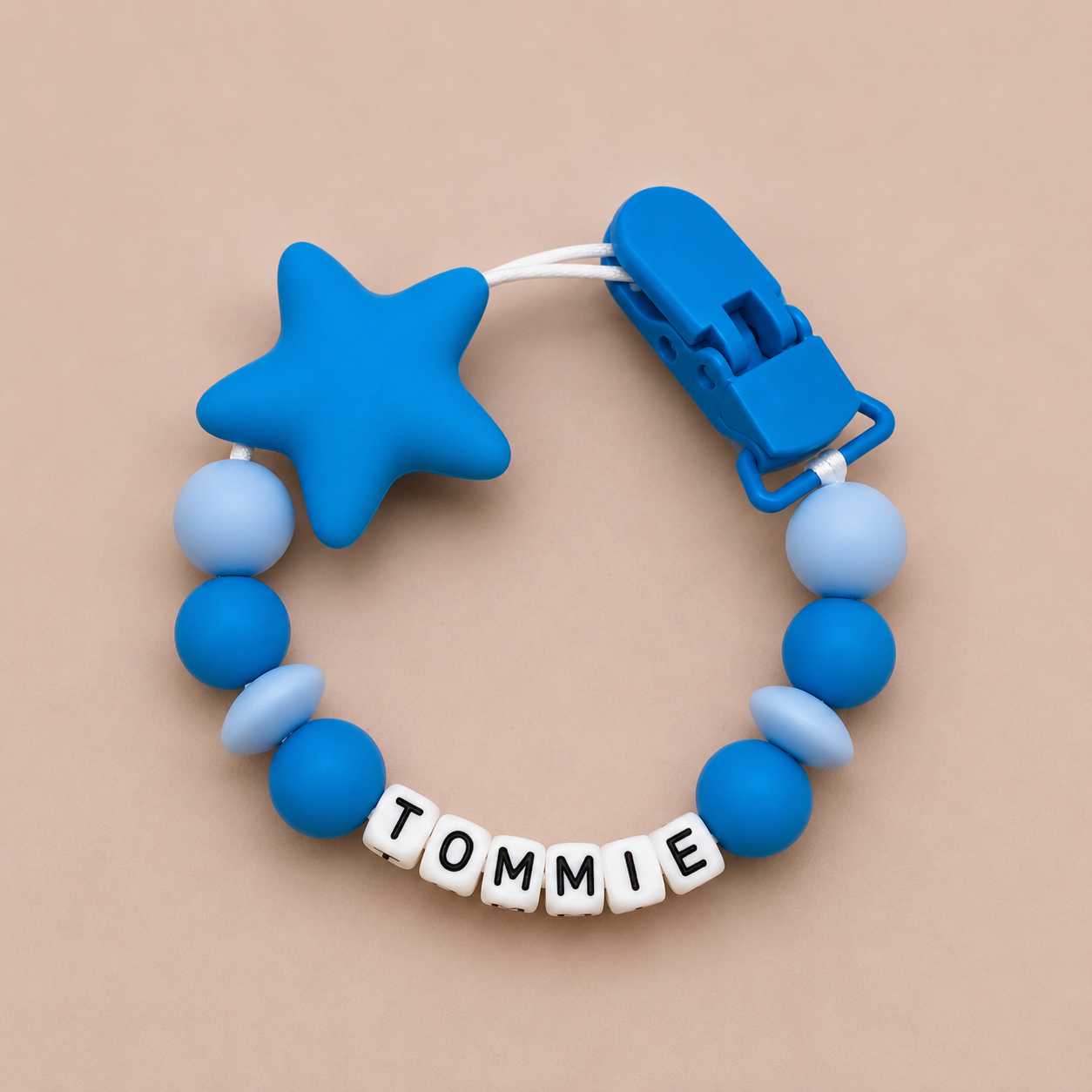 Blue dummy clip with star-shaped chewable and 'TOMMIE' name tag on a beige background