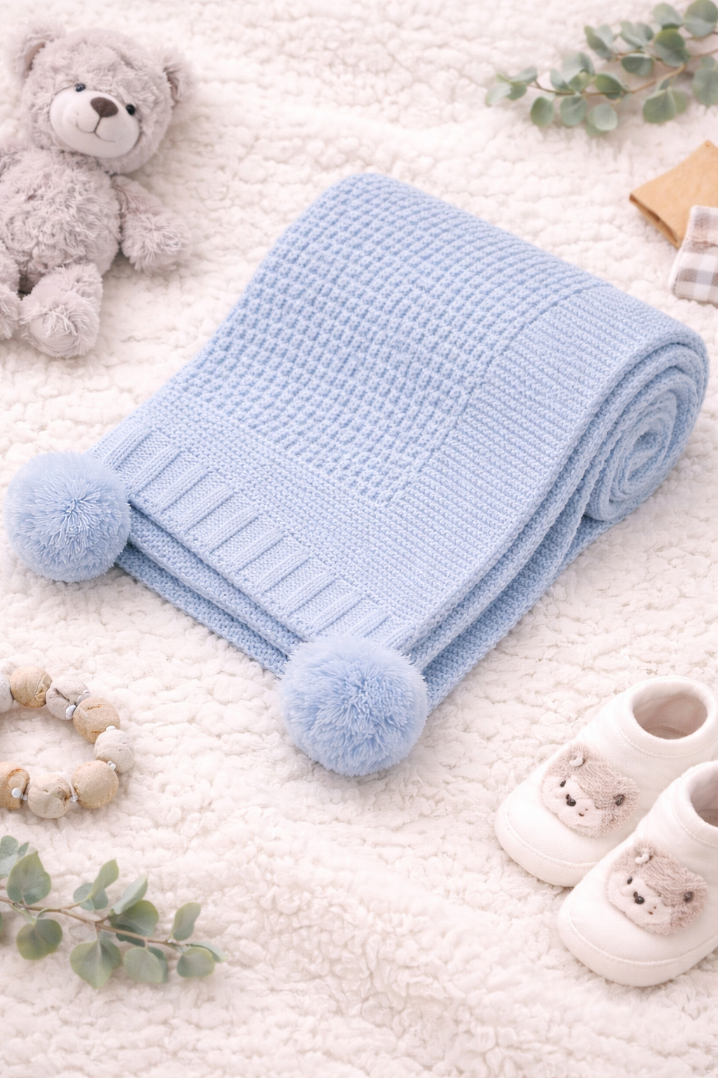 A peaceful newborn baby sleeping on a soft cream blanket, wrapped in a light blue knitted baby blanket with textured weave and fluffy pom pom corners, styled with neutral baby accessories for a cosy nursery look.