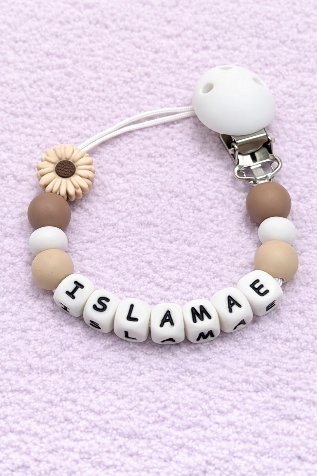 Pacifier clip with letter beads spelling 'ISLAMAE' on a light purple textured background
