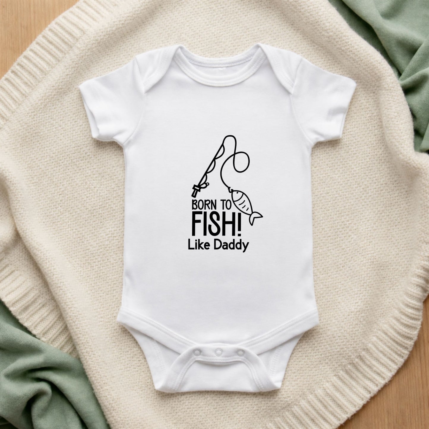 White baby onesie with 'Born to Fish! Like Daddy' text on a textured surface.