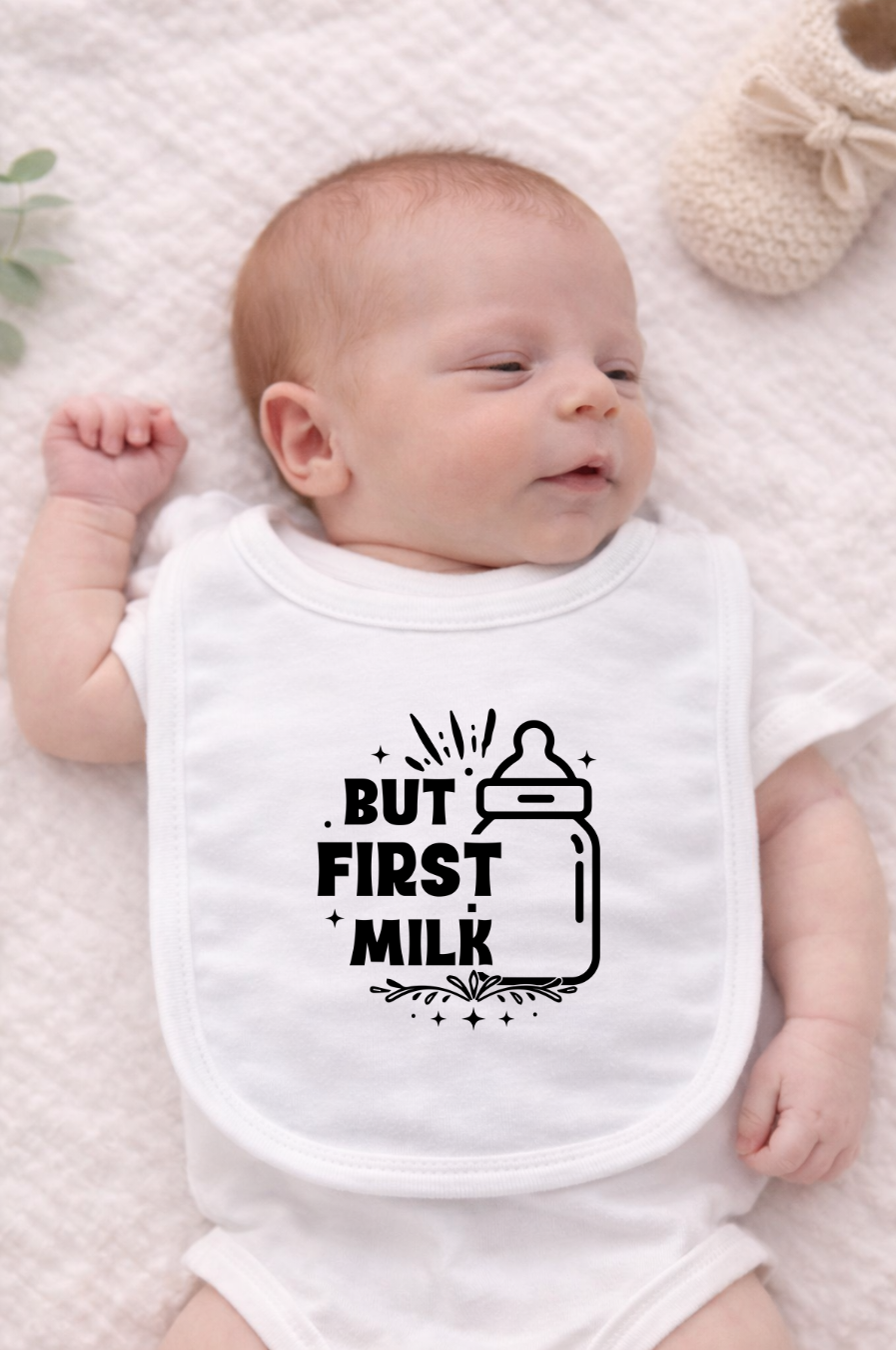 Newborn baby lying on a neutral blanket wearing a white bib with black “But First Milk” text and bottle illustration.