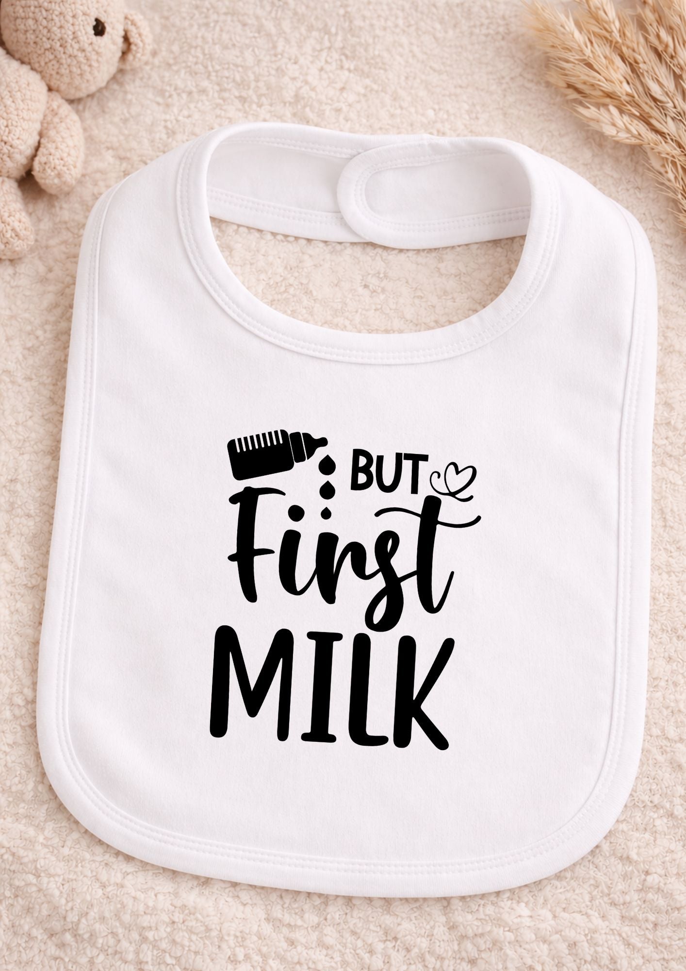 White baby bib with 'But First Milk' text on a beige surface