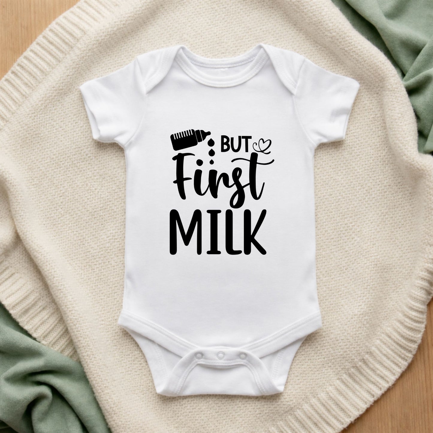 White baby onesie with 'But First Milk' text on a textured surface