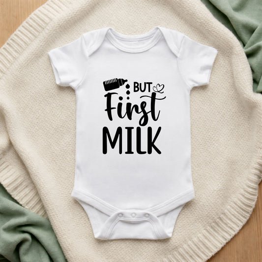 White baby onesie with 'But First Milk' text on a textured surface