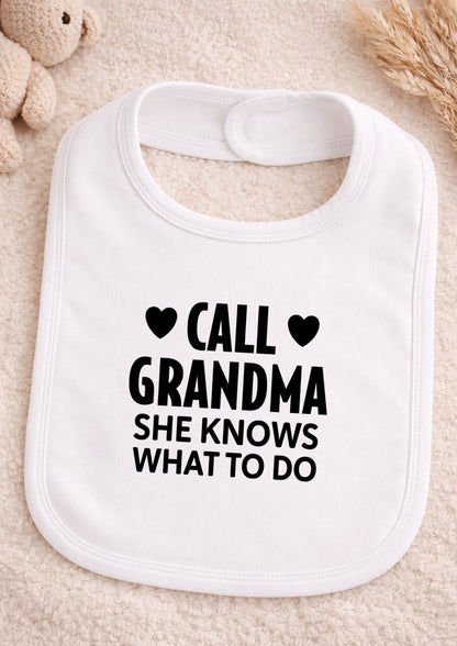 White baby bib with black text 'CALL GRANDMA SHE KNOWS WHAT TO DO' on a beige surface.