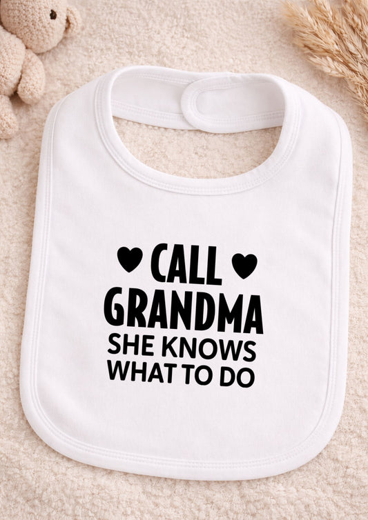 White baby bib with black text 'CALL GRANDMA SHE KNOWS WHAT TO DO' on a beige surface.