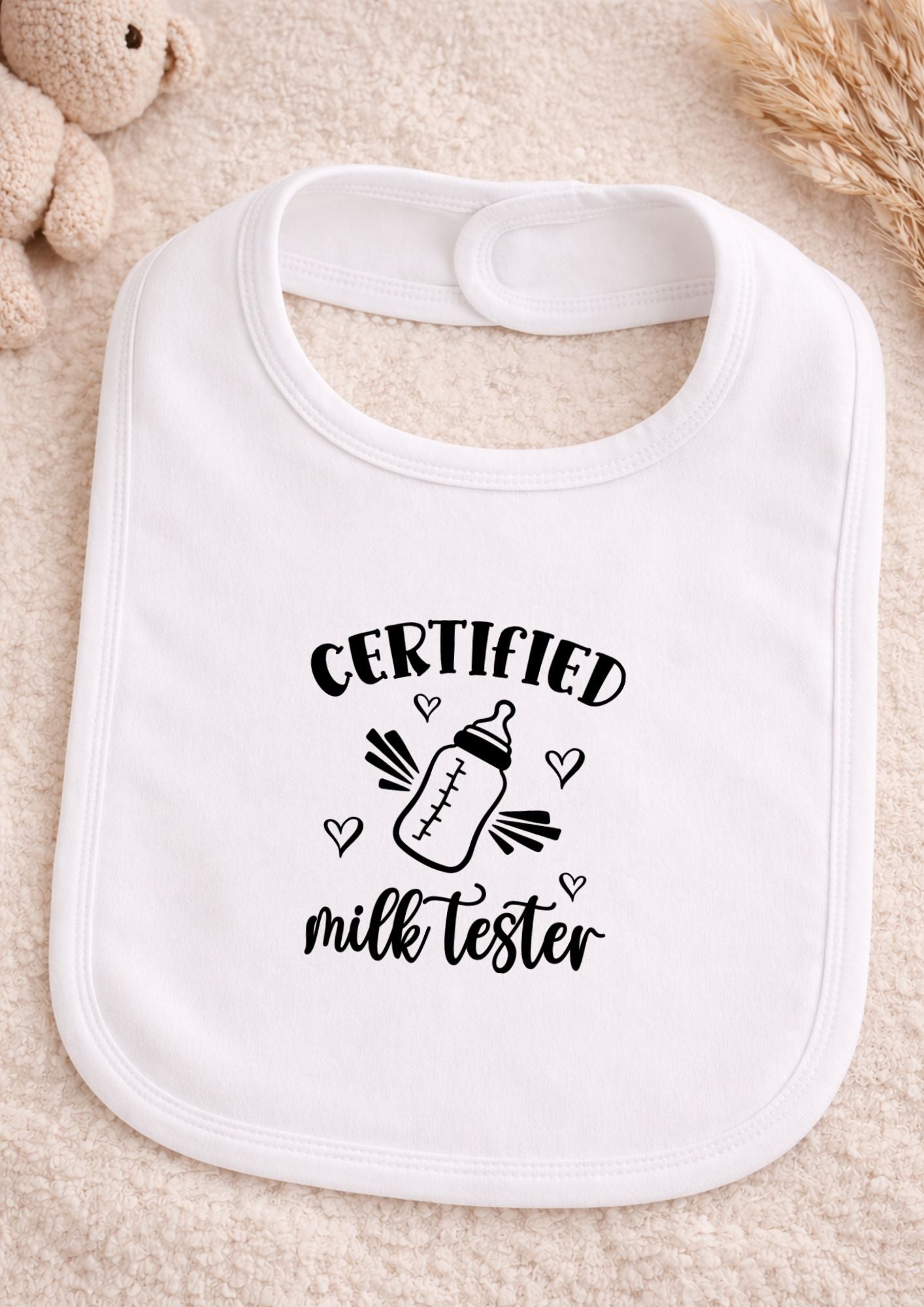 White baby bib with 'Certified Milk Tester' text and bottle graphic on a soft beige background