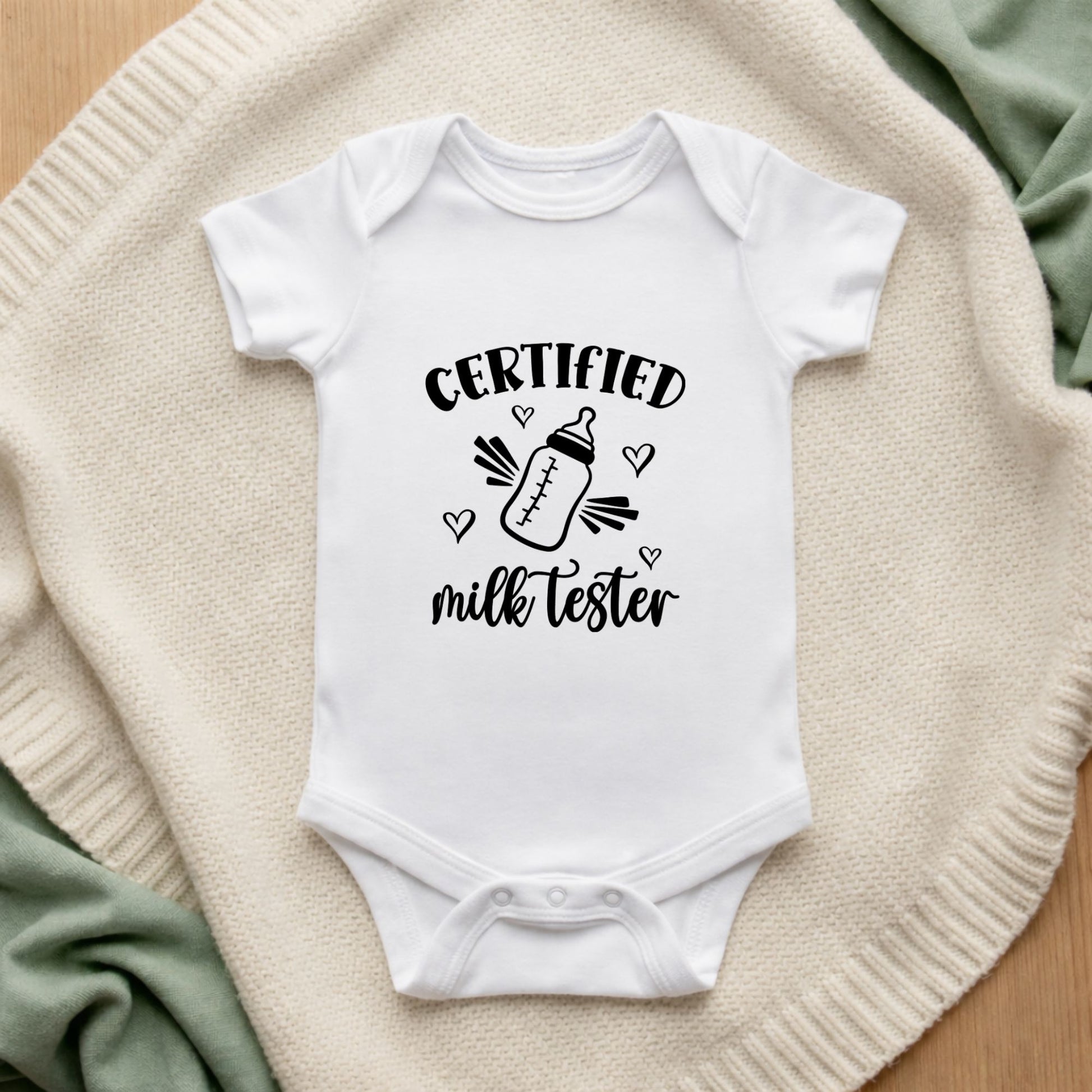 White baby onesie with 'Certified milk tester' text on a textured surface