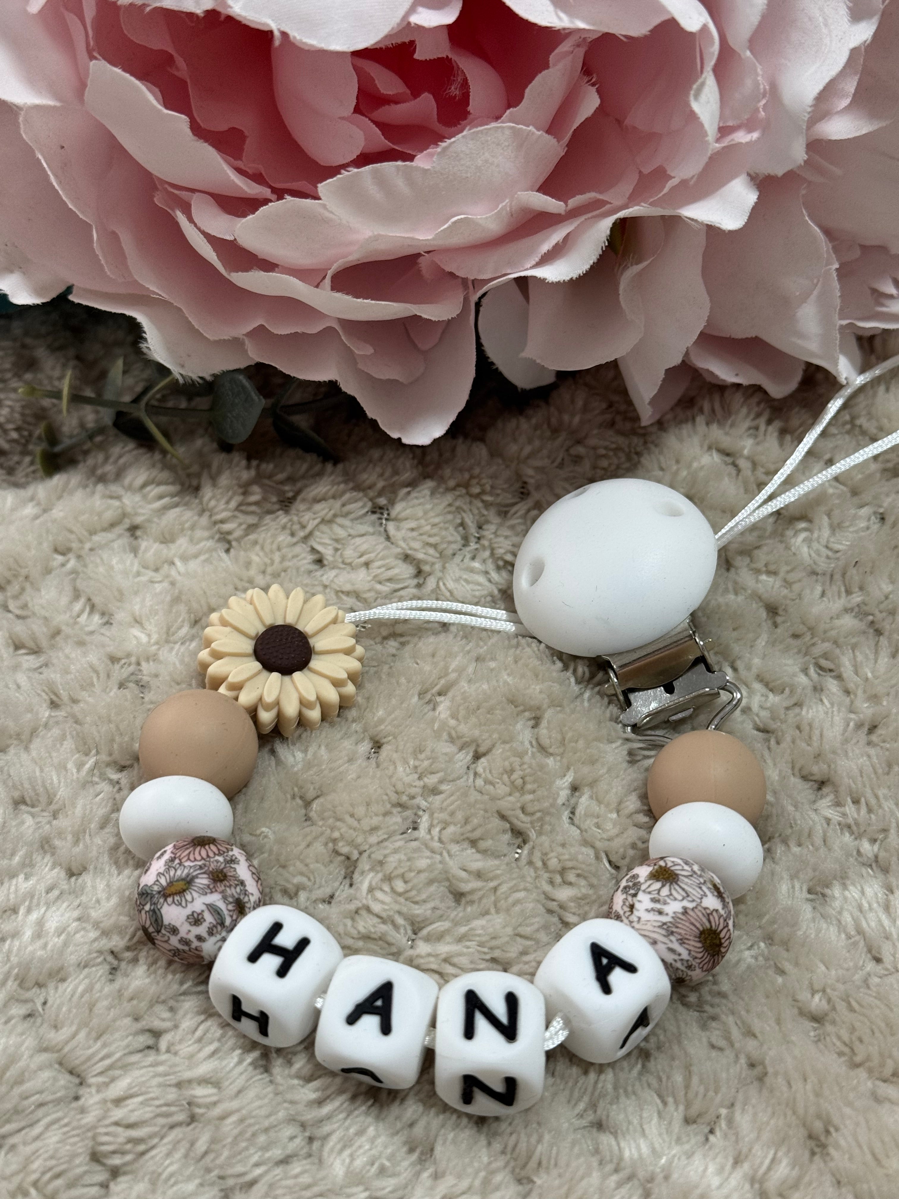Baby dummy clip with beads and letters next to a pink flower on a textured surface